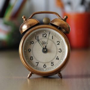 A vintage copper alarm clock with bells placed on a desk with blurred objects in the background.